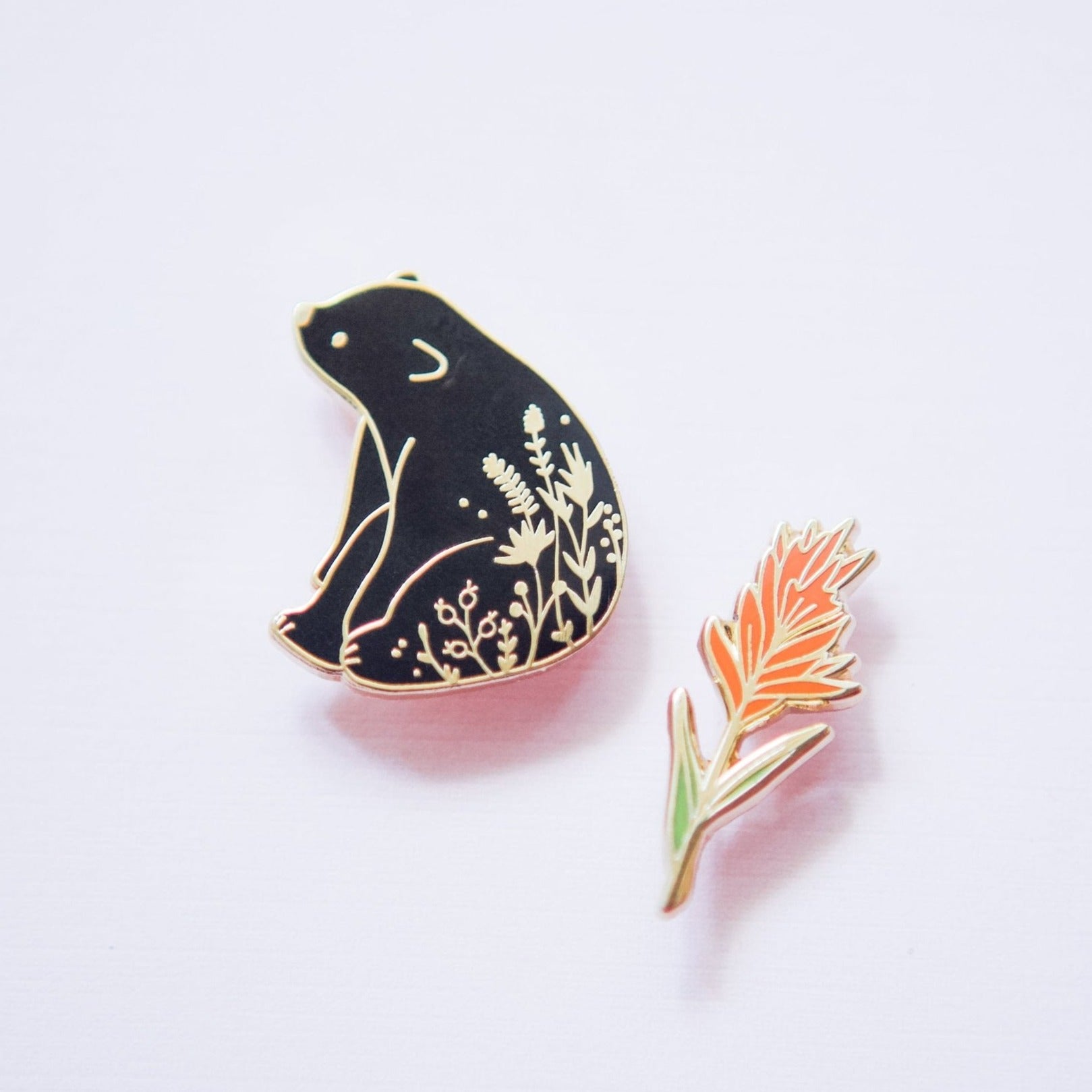 a hard enamel pin of a black bear sitting in a field of wildflowers. Enamel pins are collectible and quality additions to backpacks, hats, totes, lapels, and jackets.