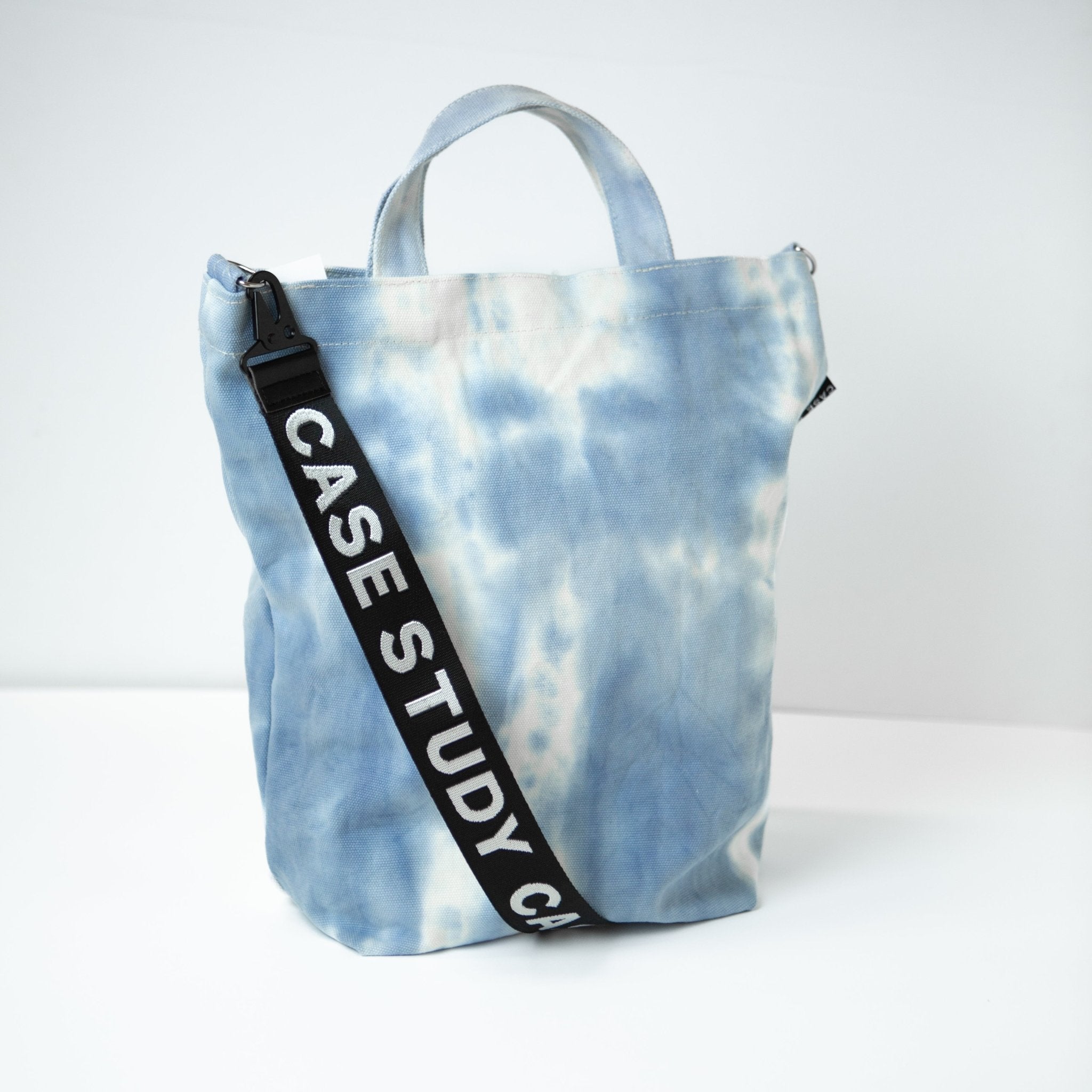 a tie dyed canvas tote in a pale blue and white color with a black and white Case Study crossbody strap