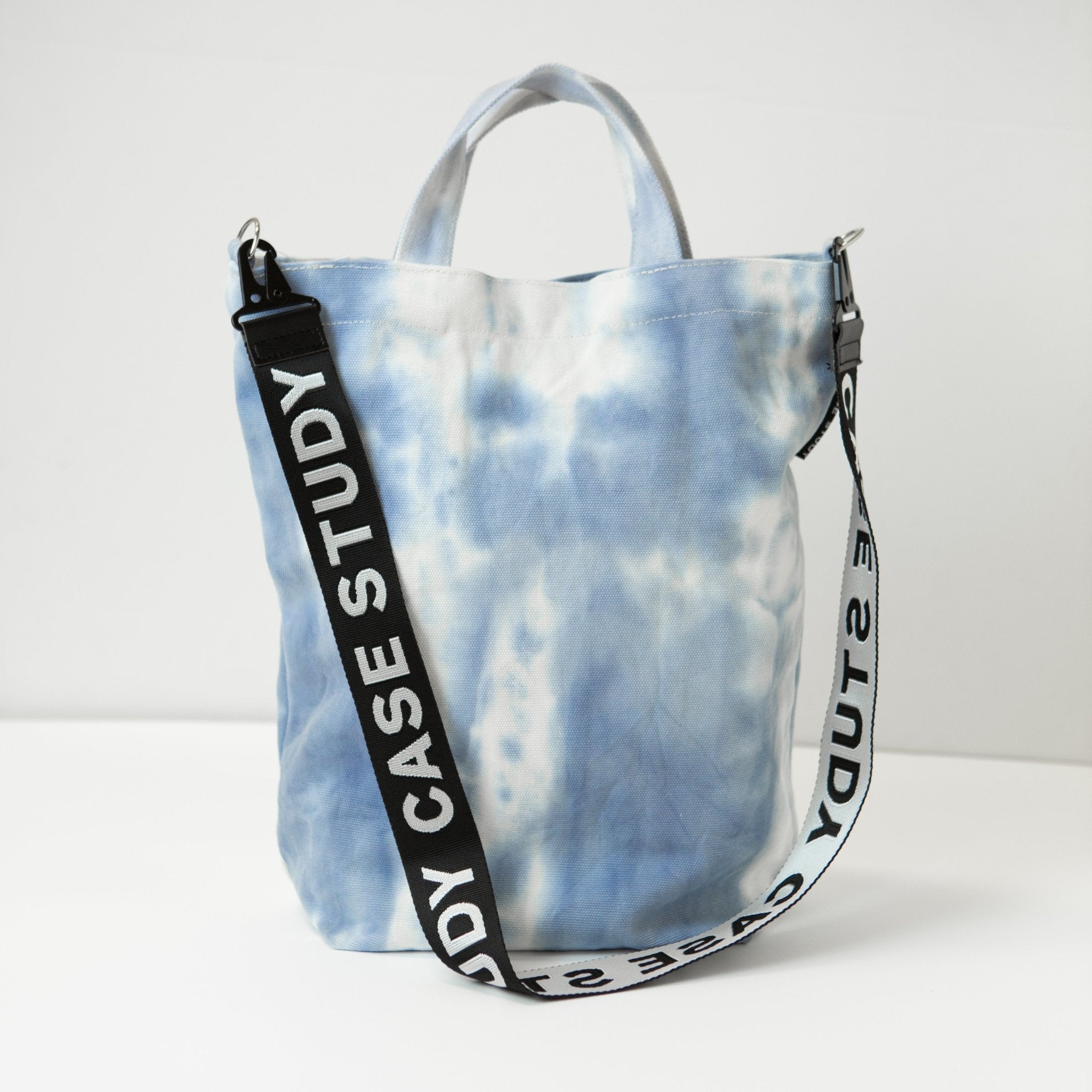a tie dyed canvas tote in a pale blue and white color with a black and white case Study crossbody strap