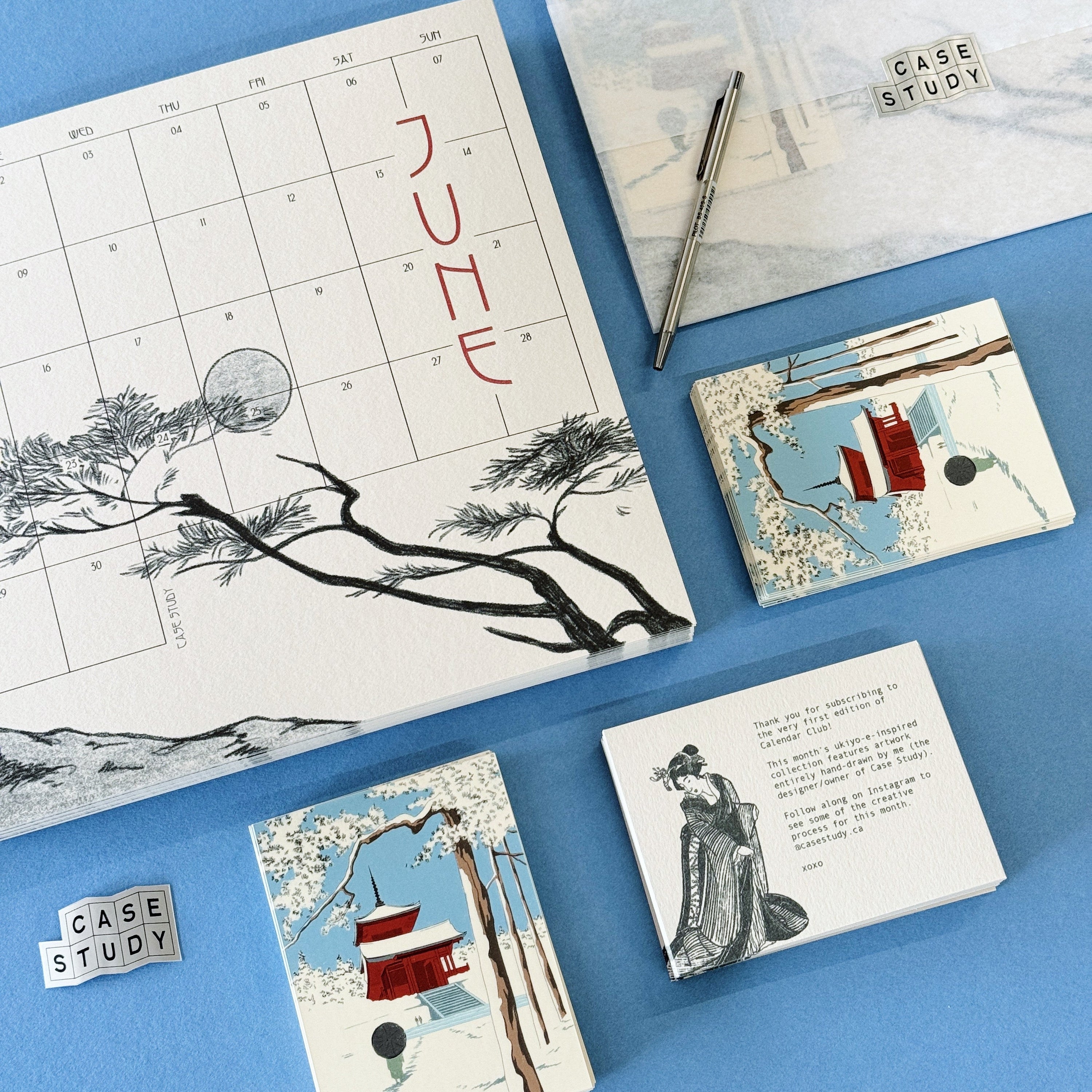 A styled flat-lay of a CASE STUDY brand stationery set on a solid blue background, featuring a June calendar page with black ink tree branches and red lettering, a pen, a notebook, and three illustrated cards.