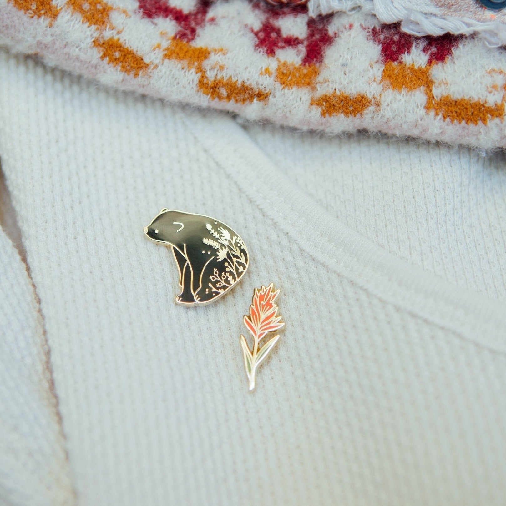 a hard enamel pin of a black bear sitting in a field of wildflowers. Enamel pins are collectible and quality additions to backpacks, hats, totes, lapels, and jackets.