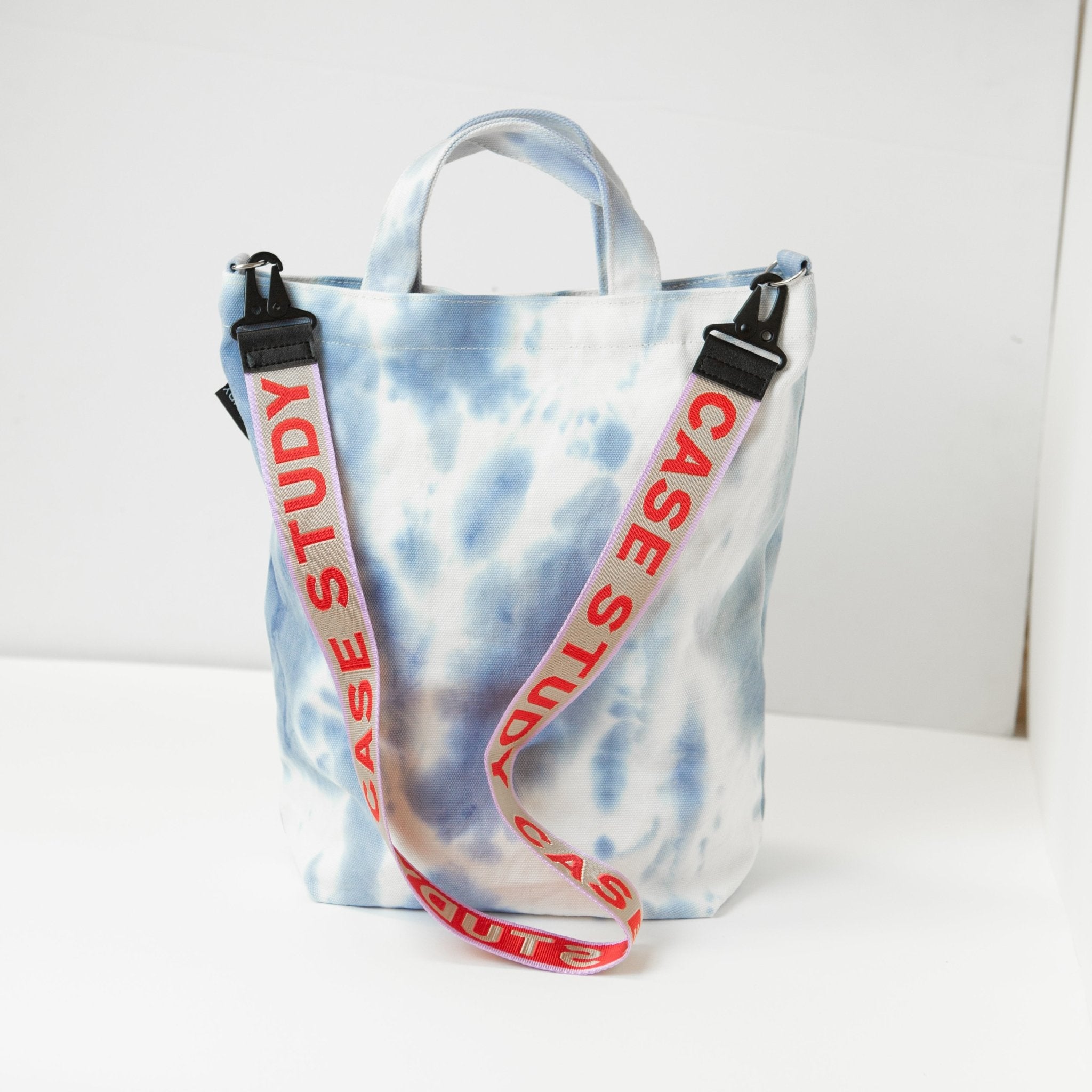 a tie dyed canvas tote in a pale blue and white color with a red and lavender Case Study crossbody strap