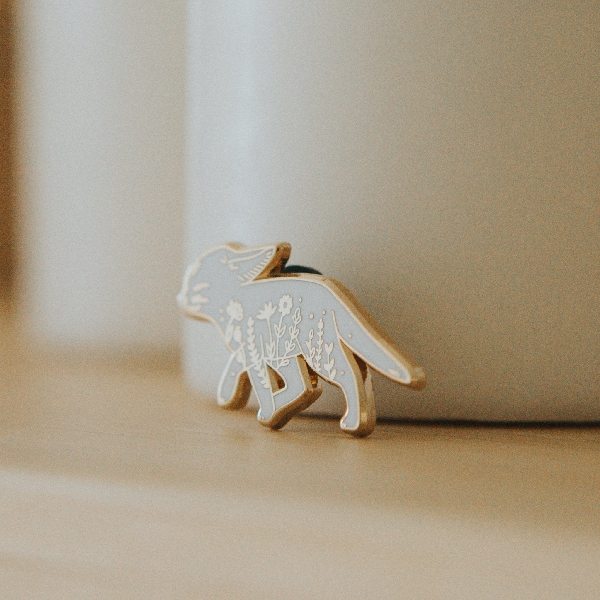 a hard enamel pin of a fennec fox and some wildflowers. Made from white hard enamel and gold metal plating